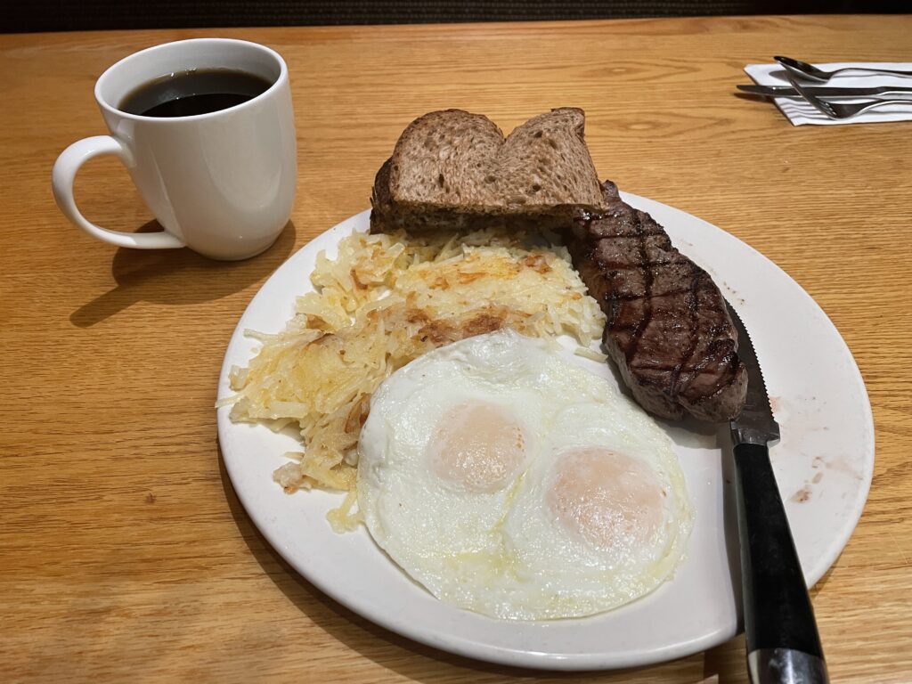 A plate with 2 eggs over easy, hash browns, toast, and a long skinny steak.