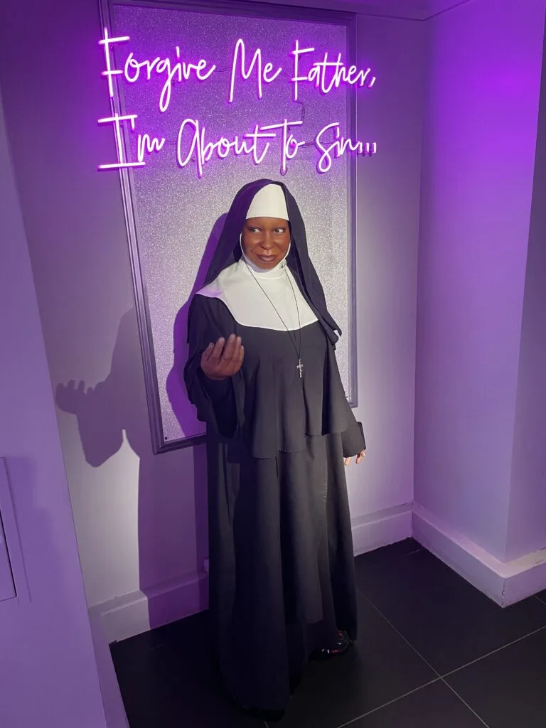 Sister Mary Clarence dressed as a nun. Above her in neon is the phrase "Forgive me father, Im about to sin".