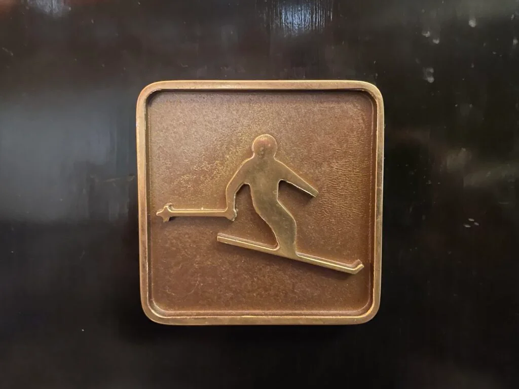 Metallic plaque with a skiier image on it which hangs in the middle of the Ski Lodge's door.