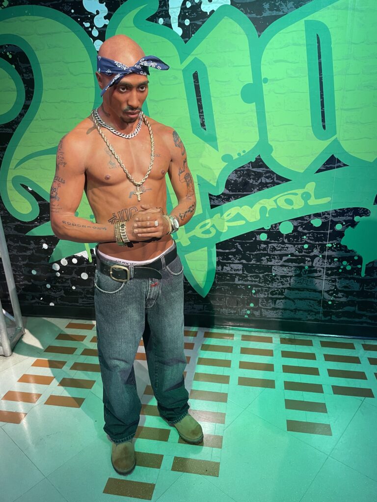 Tupac stands shirtless in front of a giant green sign that says "2Pac".