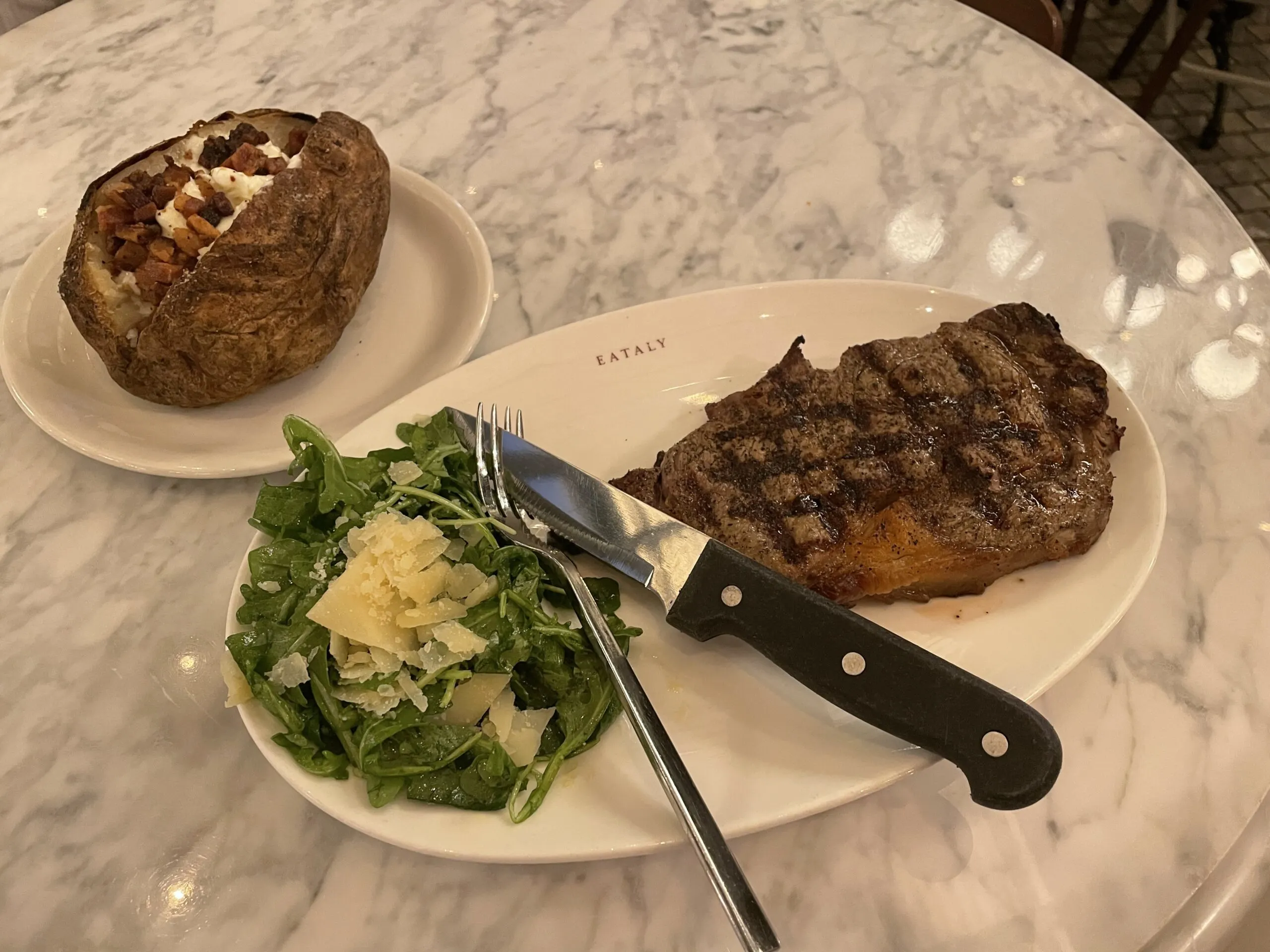 A plate with steak and a salad. On a separate plate is a massive, cheese filled baked potato.