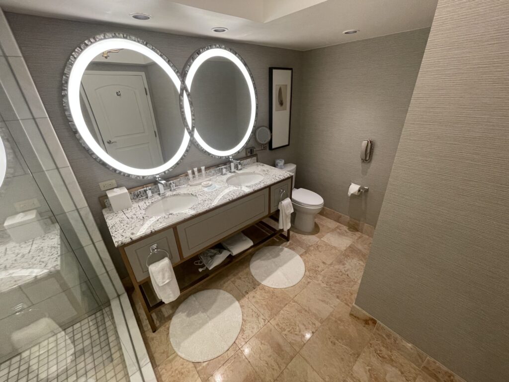 A toilet with a small phone on the wall to the right of the dual sink vanity.