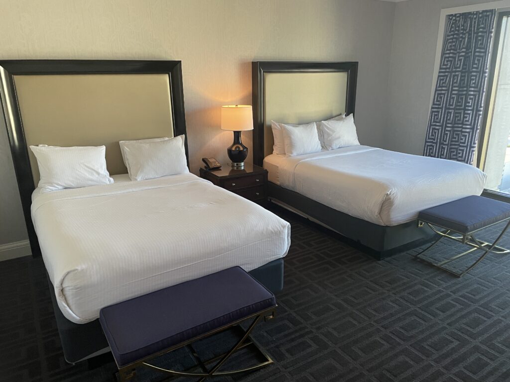 Two queeen beds with an end table between them that has an illuminated lamp atop it. 