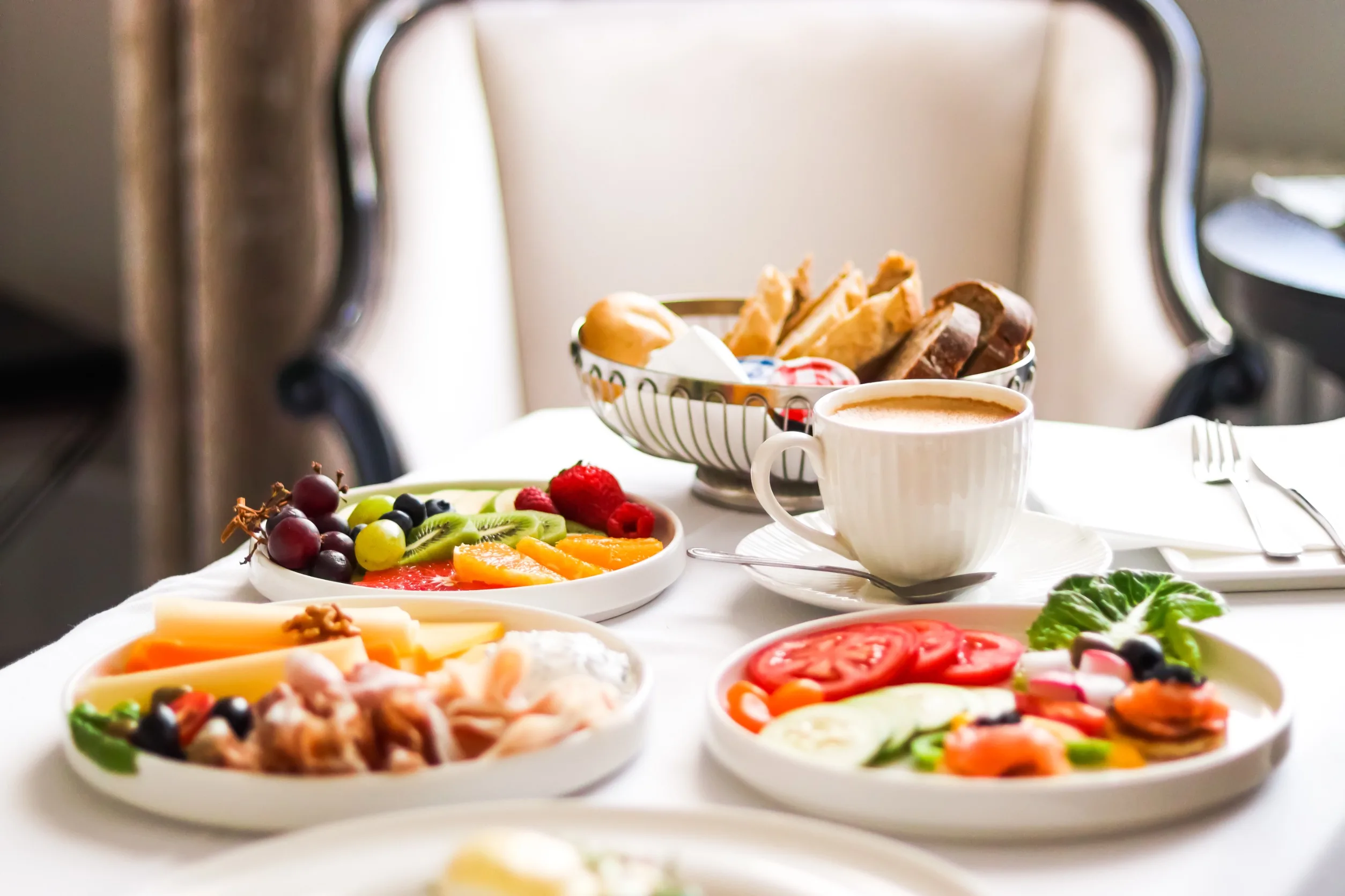 Plates of fruits and vegetables sit in front of a full cup of coffee and a bowl of assorted bread products on a table in a hotel room.