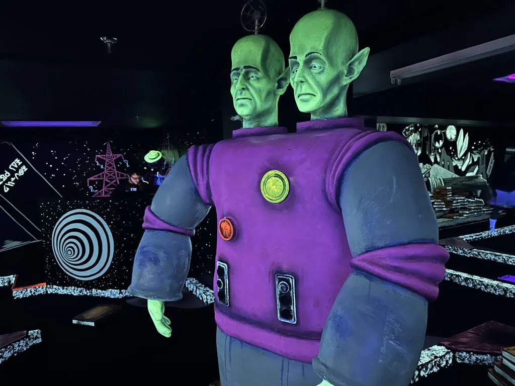 A green alien creature with two heads, wearing a purple space suit stands guard over a hole. 