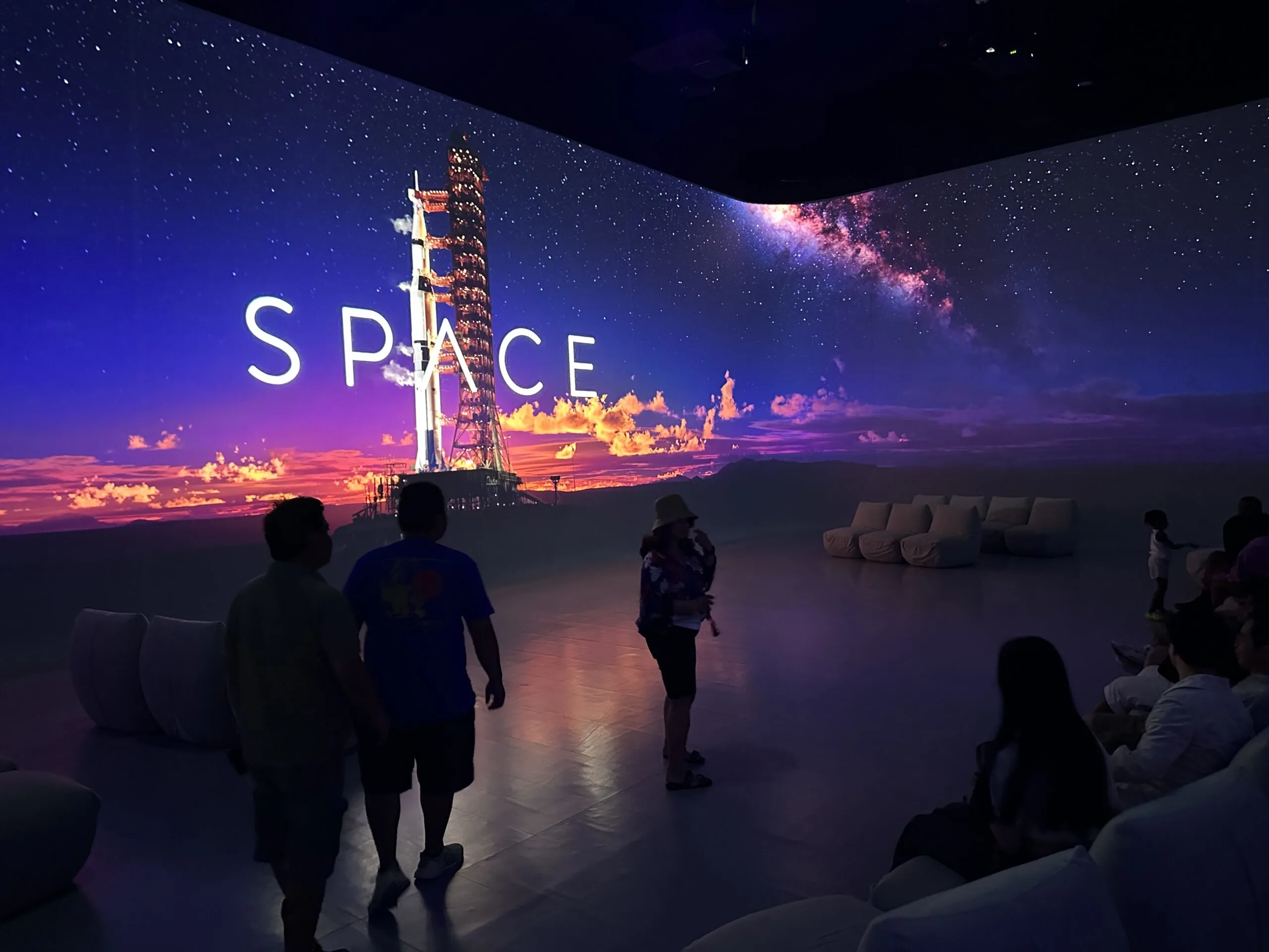 Video screen on the wall illuminates an otherwise dark room. On the screen, a space shuttle ready to take off is pictured along with the word 