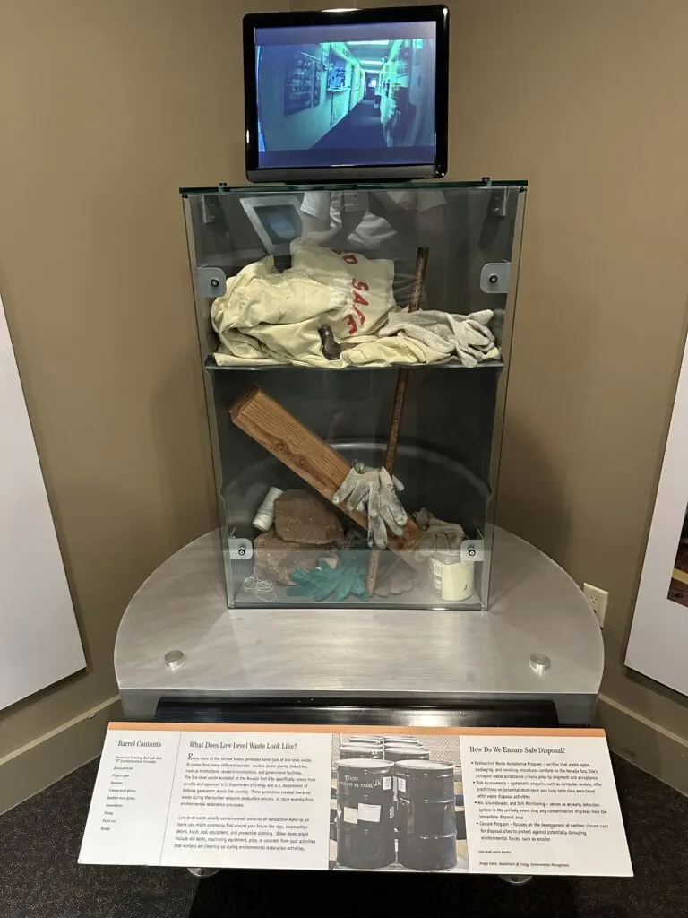 A cross section of a barrel cut in half from top to bottom. In the barrel are examples of low level radioactive watste that includes clothing, tools, and other common items. 