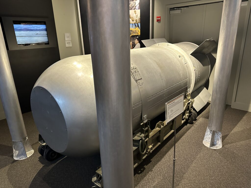 A large cylindrical silver colered bomb casing with 4 fins on the tail end. The bomb casing is sitting on a wheeled cart. 