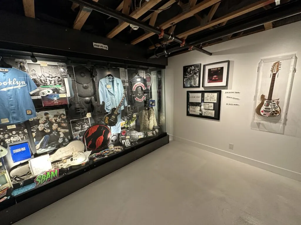 Artifacts from the pop punk era behind glass that includes guitars, symbols, clothing, and pictures. 
