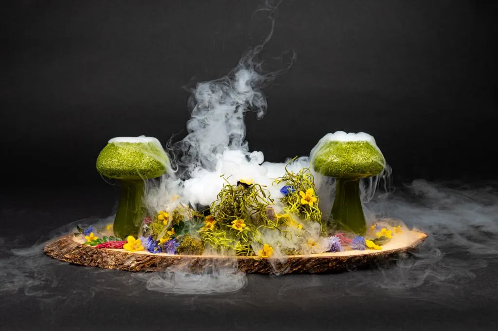 Two mushroom shaped glasses served on a wooden plate full of flowers.  Smoke is eminating from both mushrooms. 