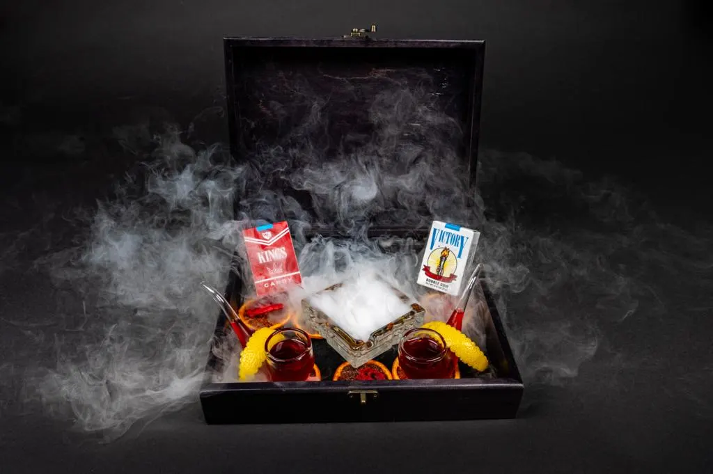 2 drinks in a cigar box that has dry ice smoke billowing out. In the back of the box are two boxes of candy cigarettes.