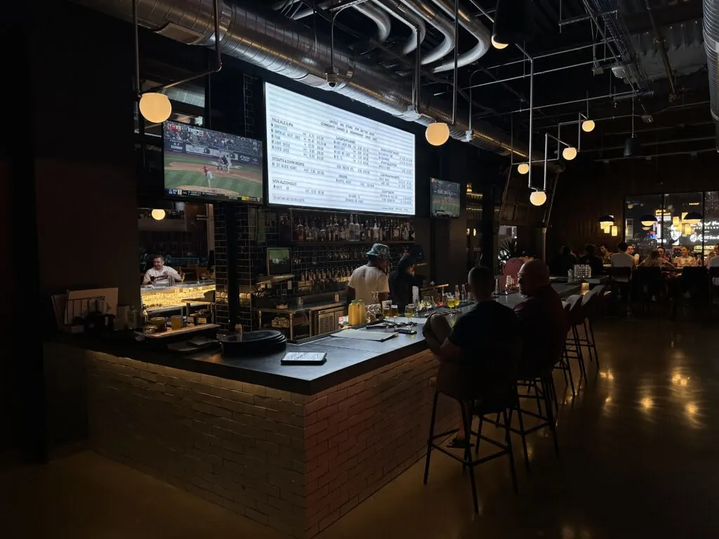 Upstairs Bar at Brewdog that has 2 tVs and an illuminated menu board on the back wall. 