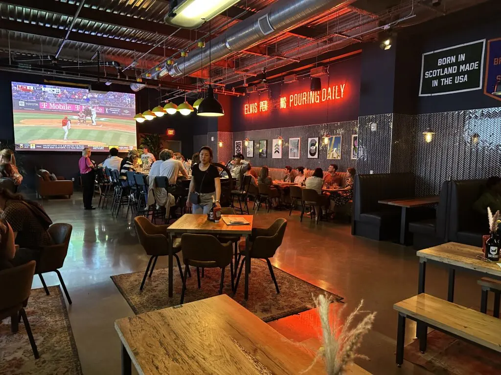 Seating area upstairs, which has tables, chairs, and a large tv in the background. 