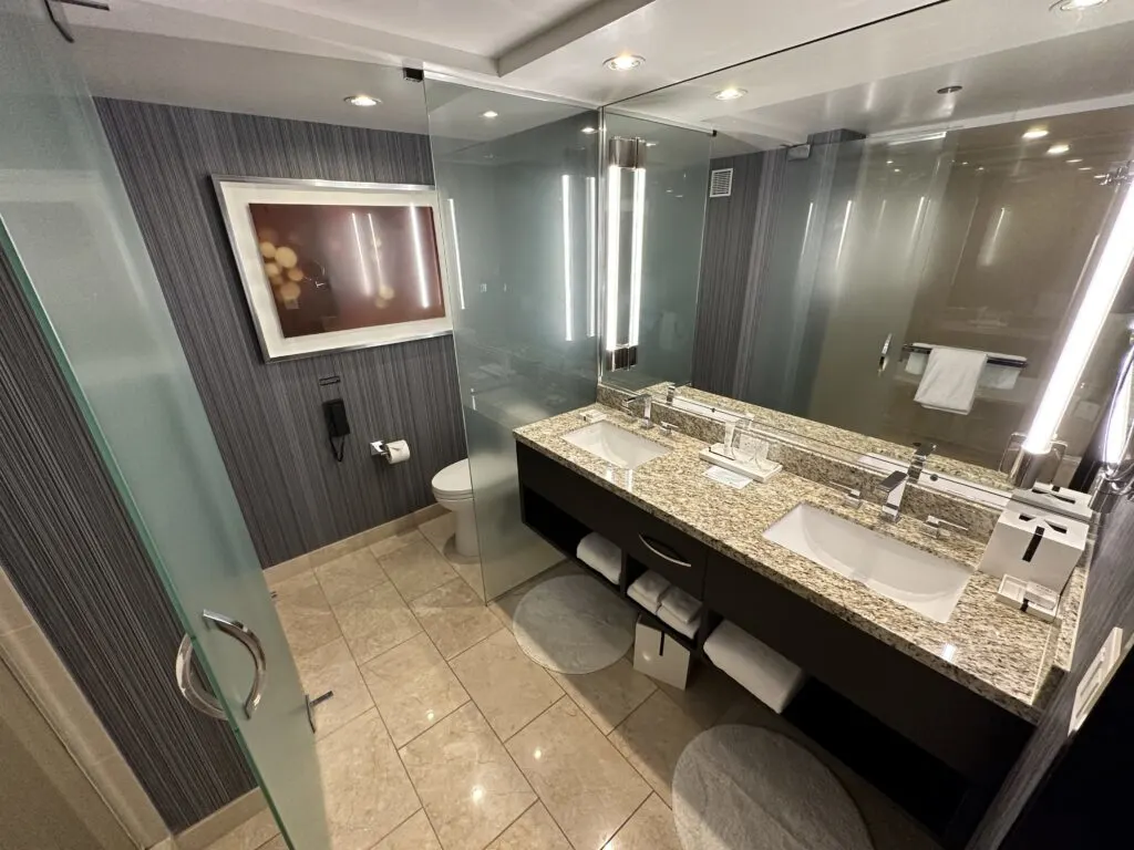 A dual vanity sink made of brownish marble with a large mirror above it with unique bar lights on either side. Behind the vanity is a small separate room where a toilet is partially visible. 