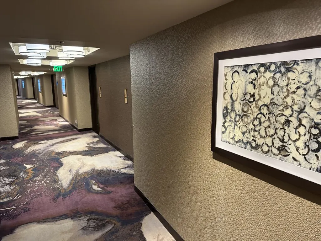 A hotel hallway at Aria with purple carpet, a painting on the wall, and decorative lighting on the ceilings. 