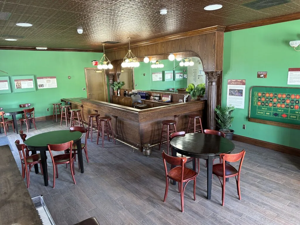 Interior of the Arizona Club Saloon which has bright green walls, a wooden bar, and tables with chairs scattered about.