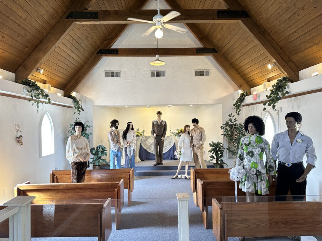 8 manequins that are made to appear to be part of a wedding ceremony are standing in the pews, looking back at the entrance. 
