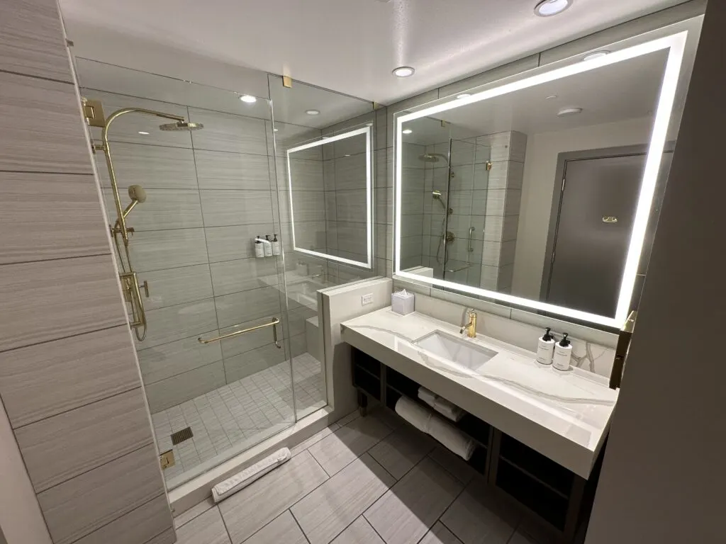 A single sink vanity topped by a large, illuminated mirror. A glass enclosed shower is located next to it. 