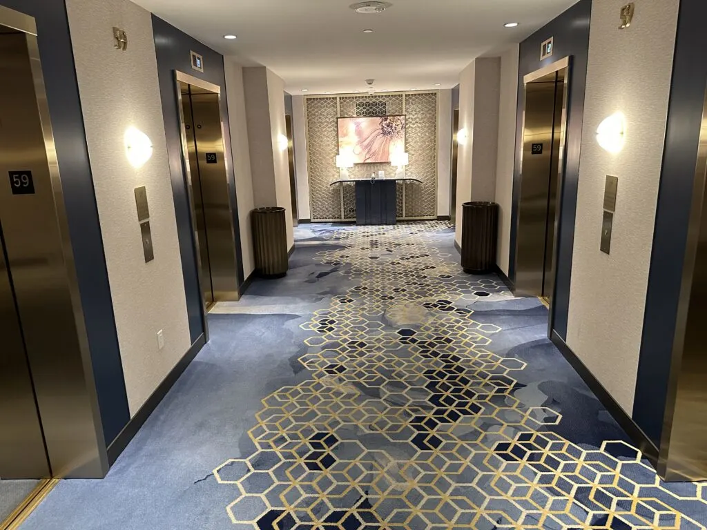 An elevator lobby on my floor at Hilton, which has blue carpet and a large mirror flanked by elevator doors on either side. 