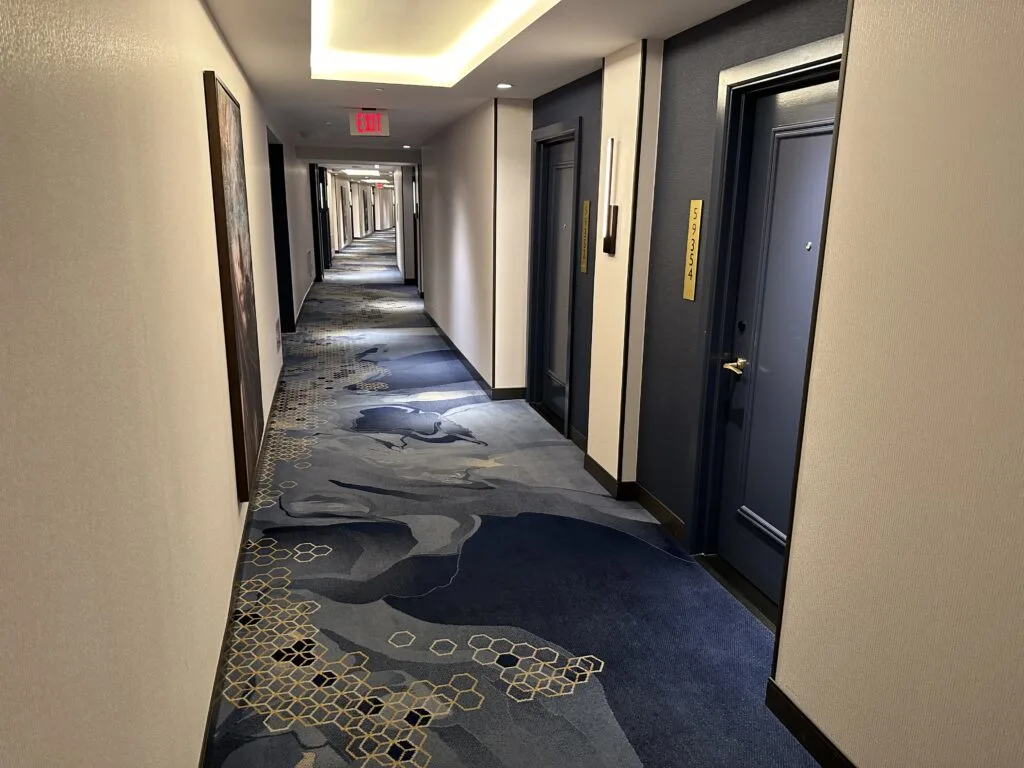 A brightly lit hotel hallway extends into the distance. The carpet is a dark blue with wavy floral patterns and the doors are a darker blue. 