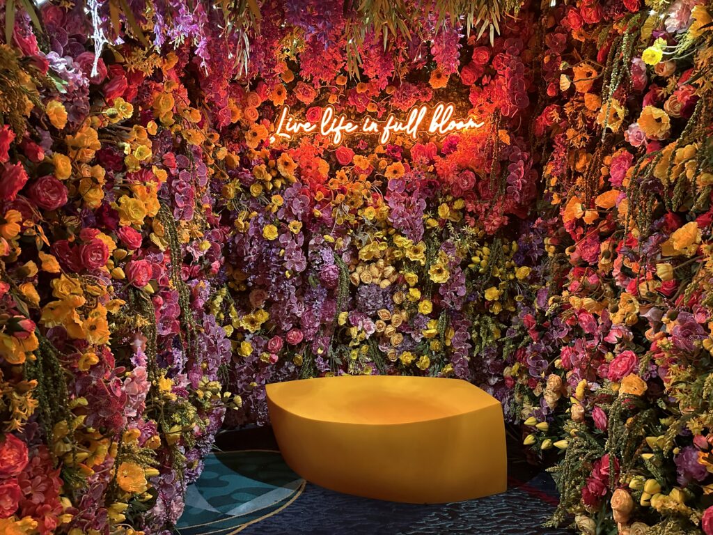 A photo opportunity with a bench surrounded by fake plants and flowers on 3 walls. On the back wall above where your head would be is a red neon sign that says "Live life in full bloom".