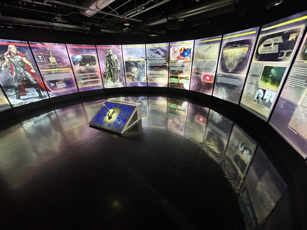 A circular room with illuminated walls full of images and character information. In the middle of the room is a large hammer. 