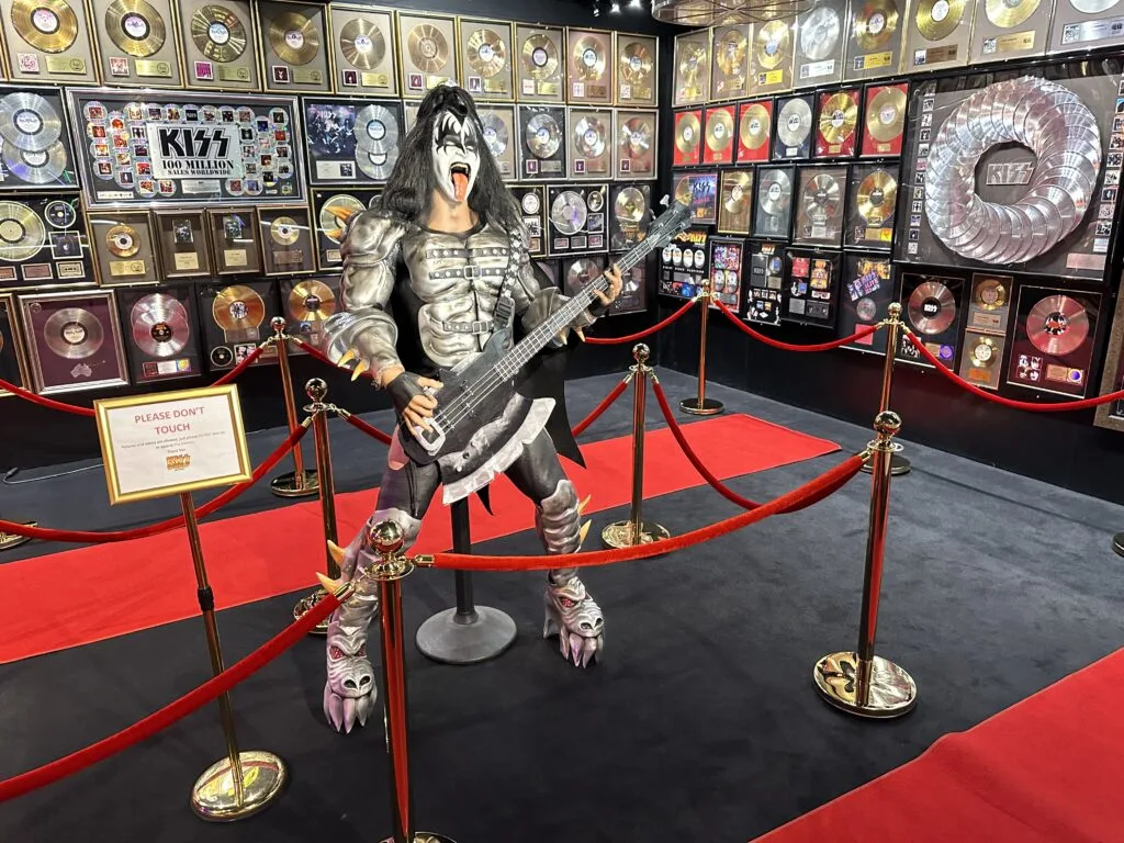 A Kiss statue plays a guitar in front of 2 walls that are full of metallic records. 