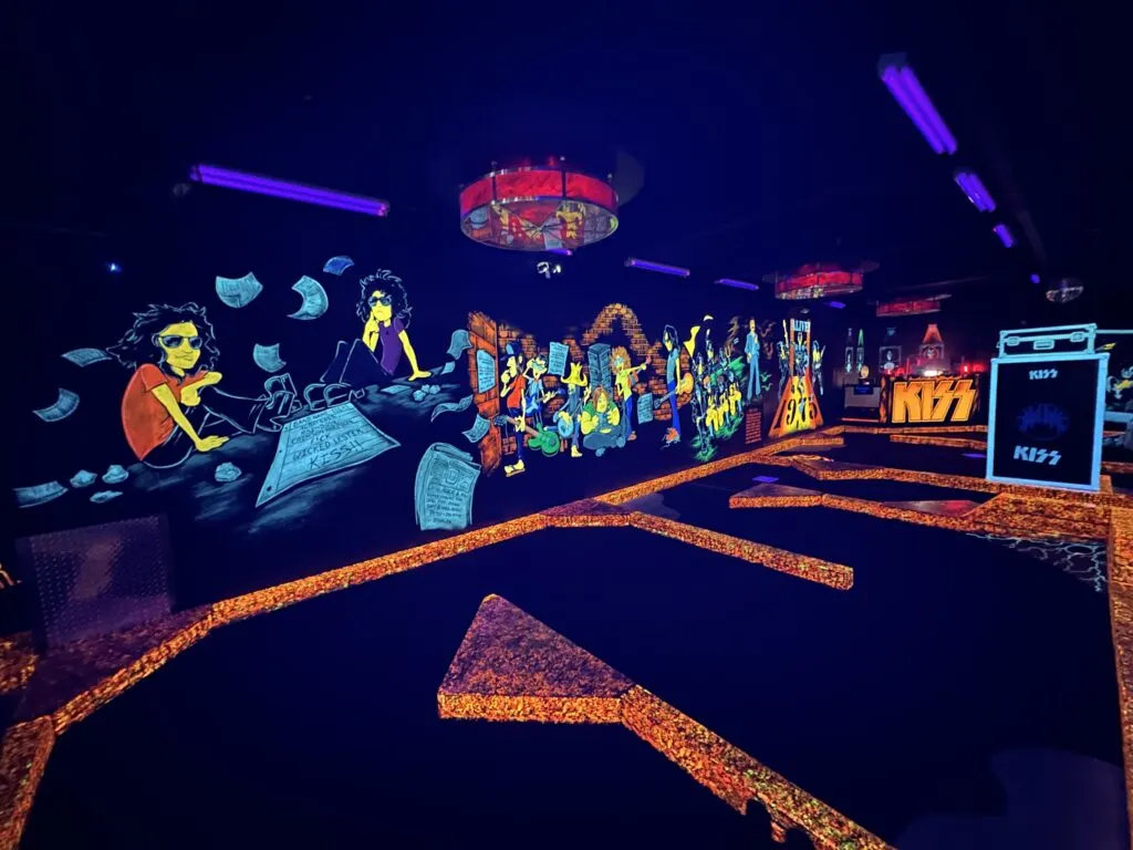 One of the walls at the mini golf course that has a large mural of band members and other influential figures glowing under the blacklighting. 