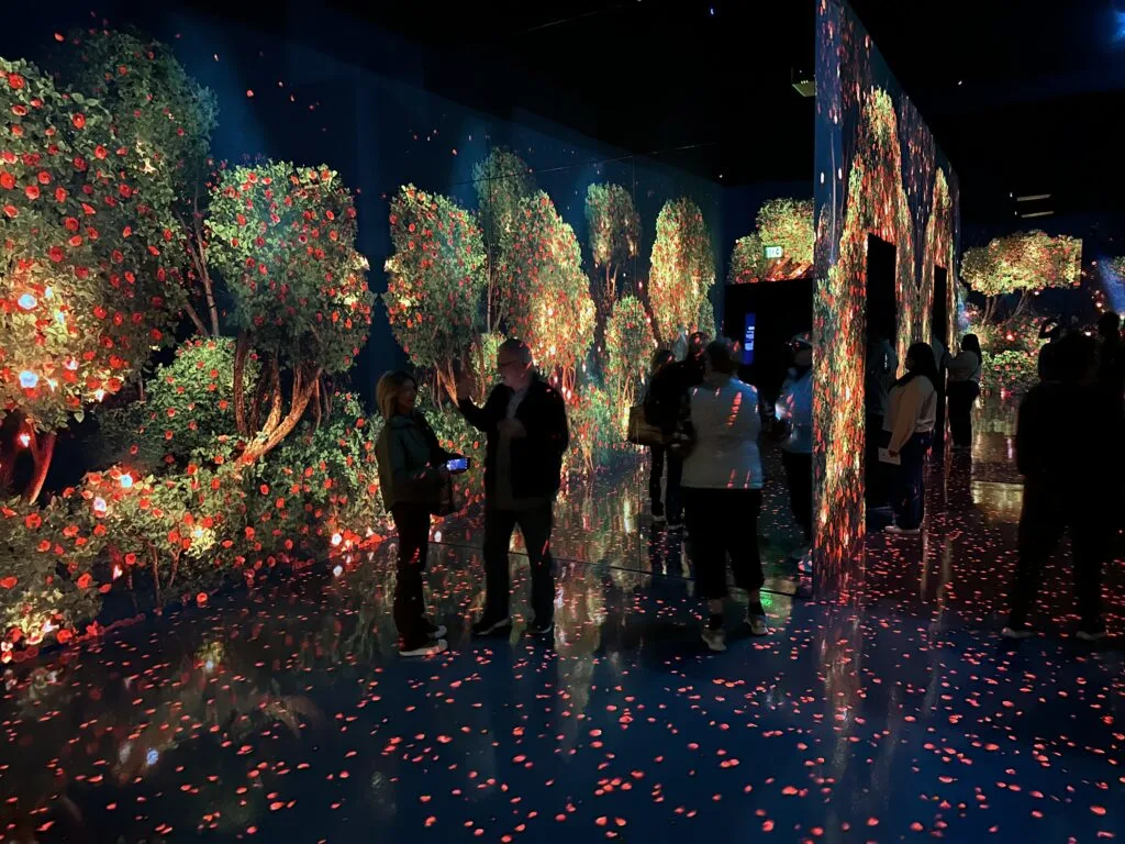 A dark room where green plants with red flowers on them cover the walls. Flower petals are projected on to the floor. 