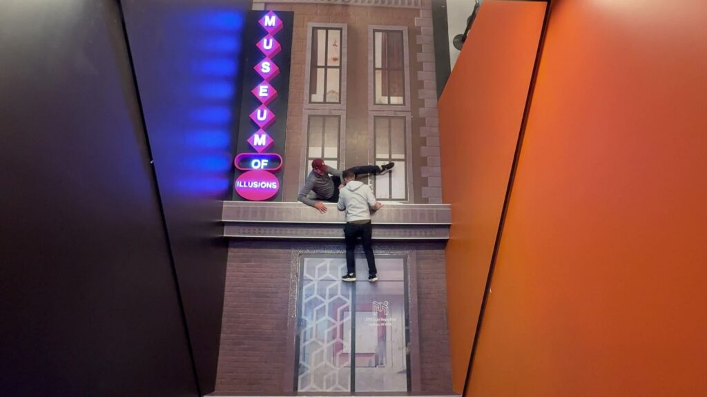 A building facade with a ledge that I am on. The illusion makes it appear my brother fella and I am pulling him back up.