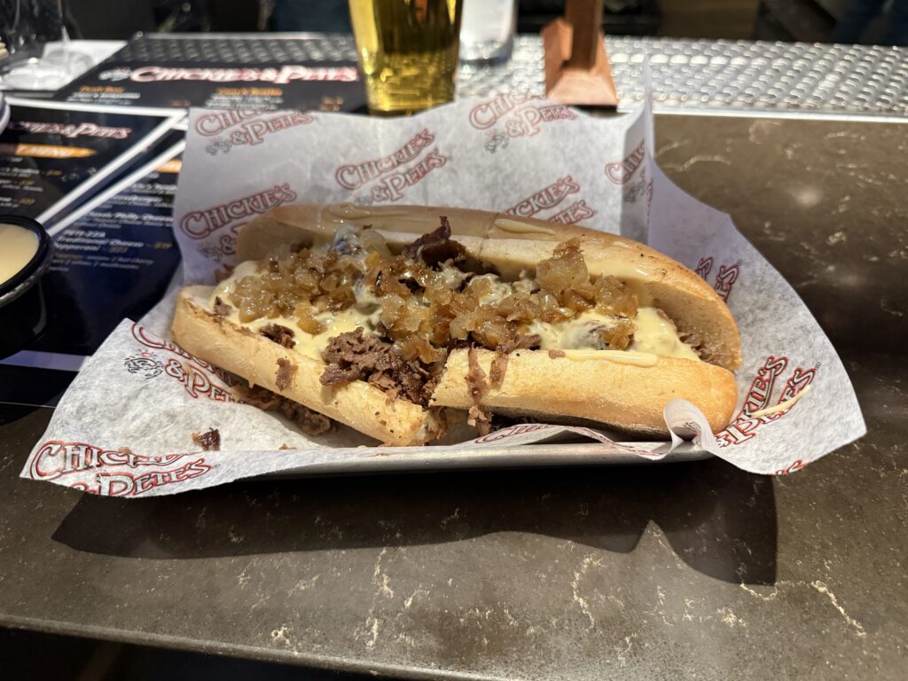 A Philly Cheesesteak in a basket, which is about a foot long and overflowing with meat and cheese. 