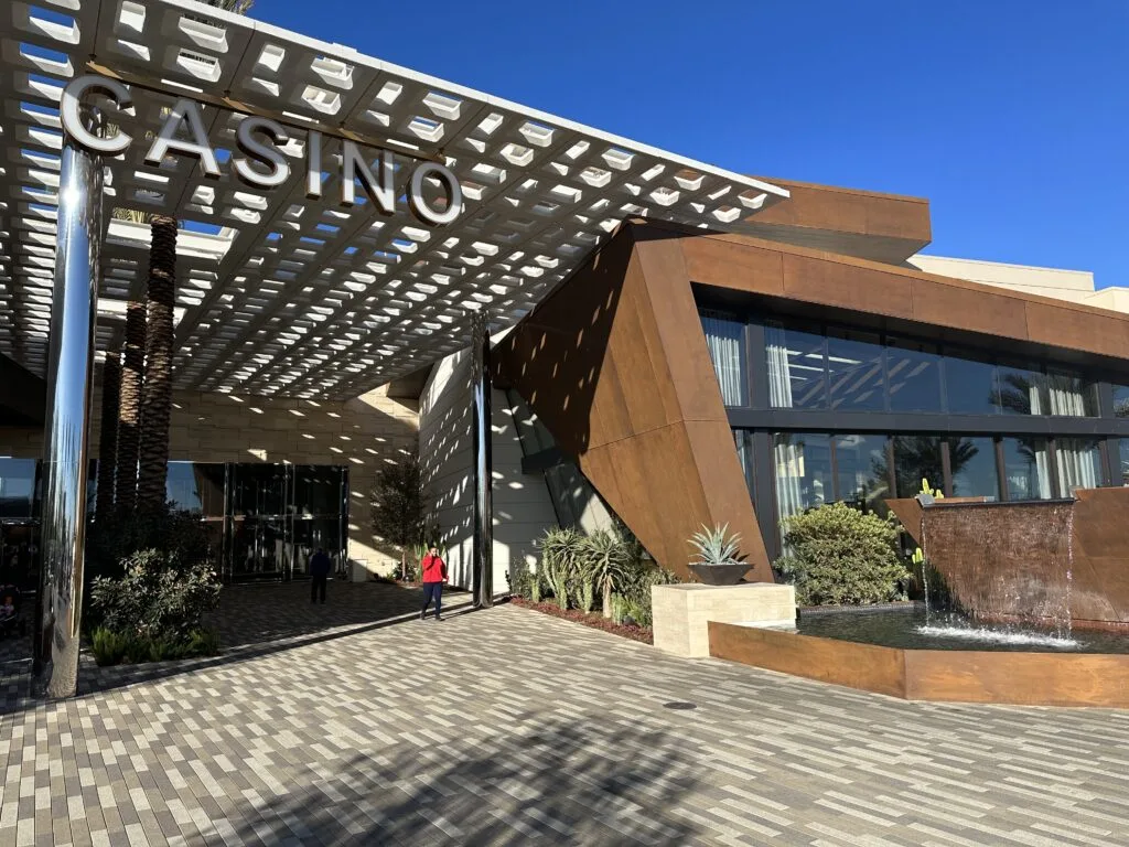Durango Las Vegas Casino Entrance flanked by plant life and a water fountain. Several tall palm trees are on the walkway in, and there are holes made in the ceiling of the entryway for them to pop through. 
