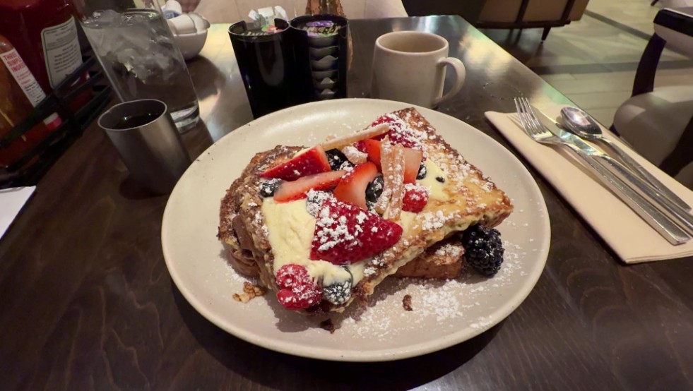 Plate of waffles topped with mixed berries (strawberries, raspberries, blueberries) and cream, dusted with powdered sugar, on a dark table in a cafe.