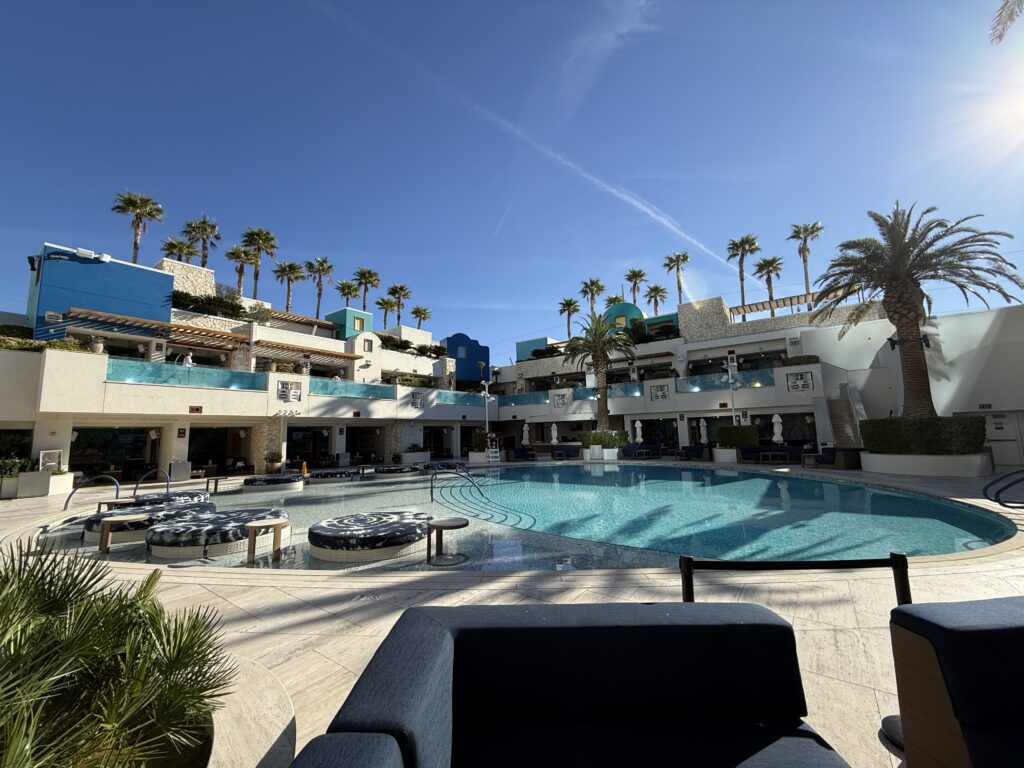 Resort poolside with turquoise water, palm trees, and white modern buildings under a bright blue sky.