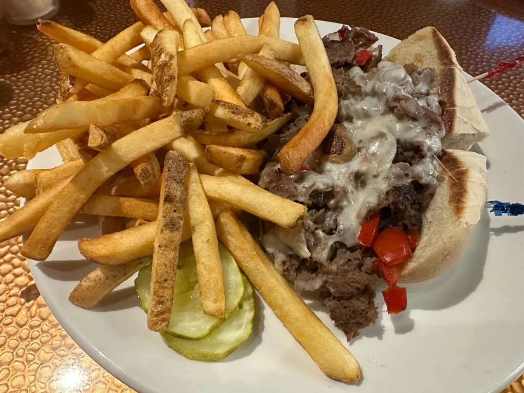 A philly cheesesteeak next to a pile of french fries and 2 pickle slices on a plate.