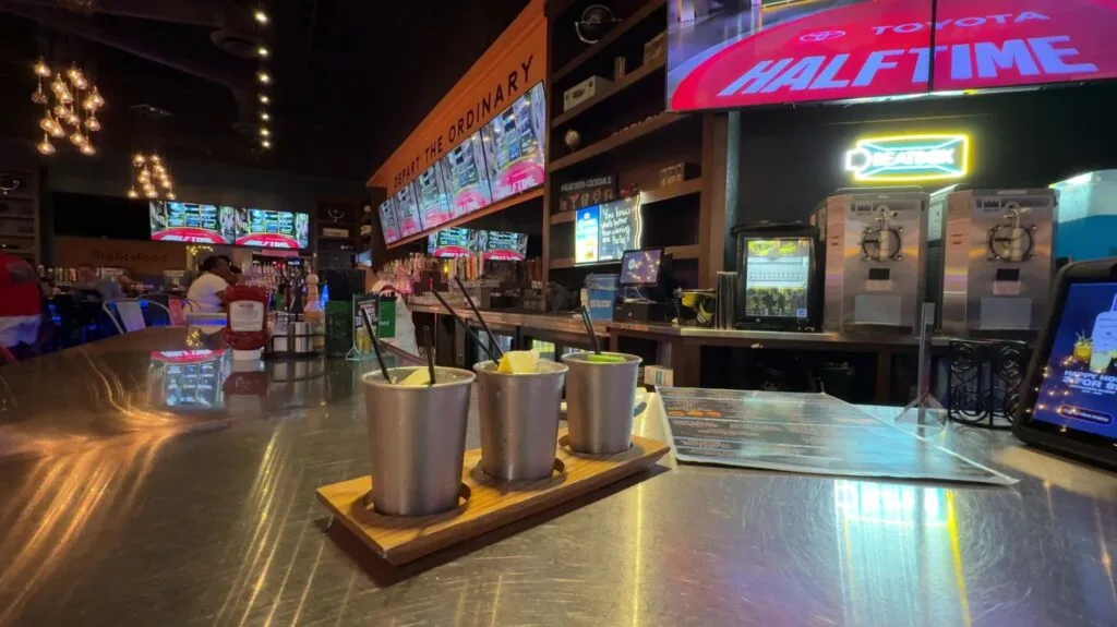 A flight of 3 drinks in metal cups with TVs showing football in the background.