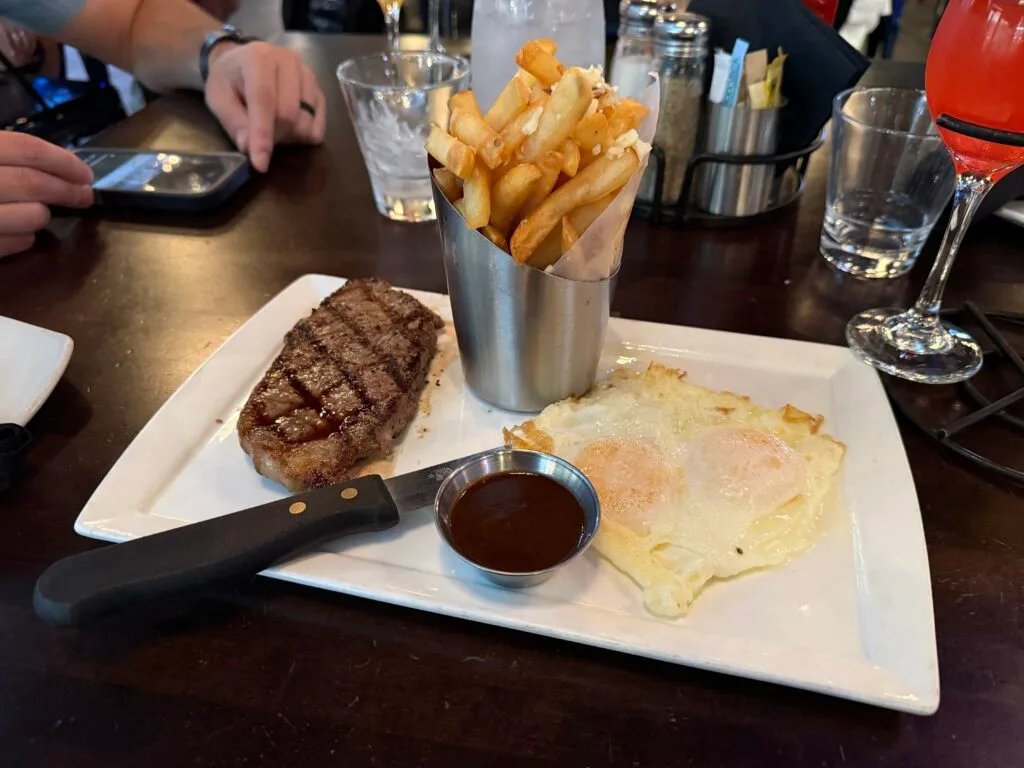 A rectangle white plate with an oval shaped steak, a basket of french fries, and two eggs over easy.