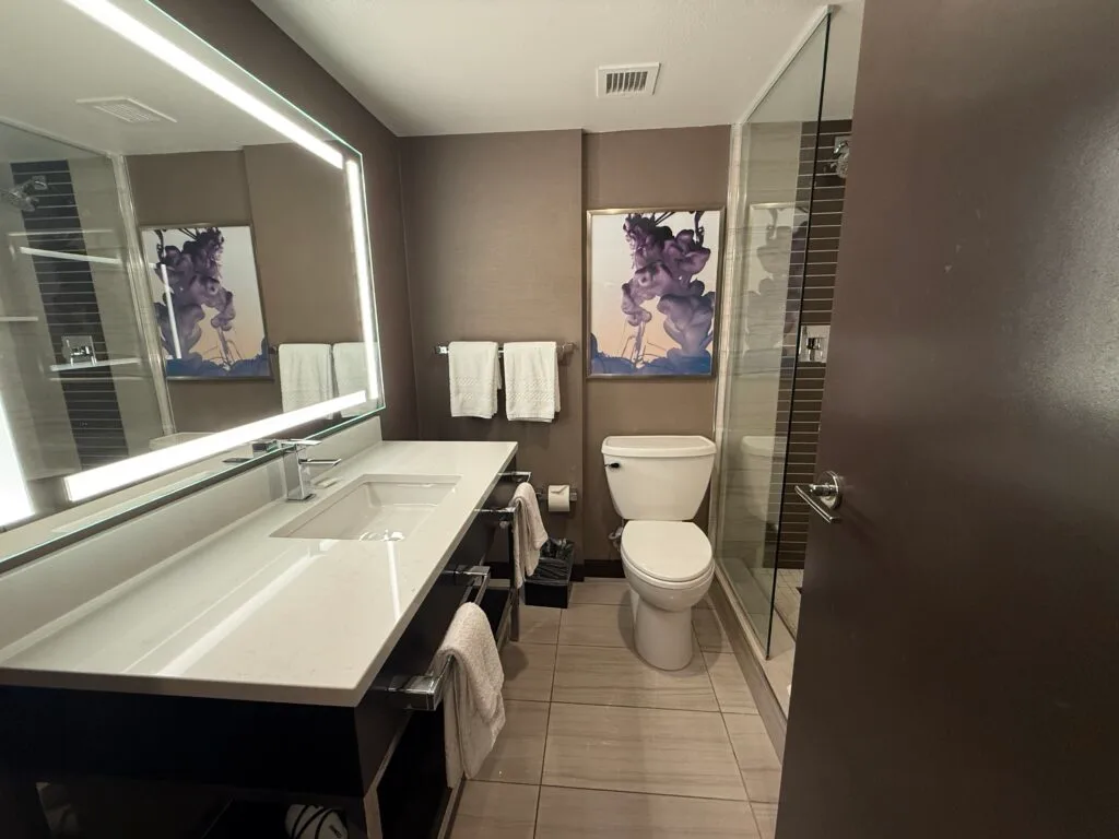 A skinny bathroom at Harrah's, with a sink, vanity, and illuminated mirror to the left, toilet straight ahead, and shower to the right.