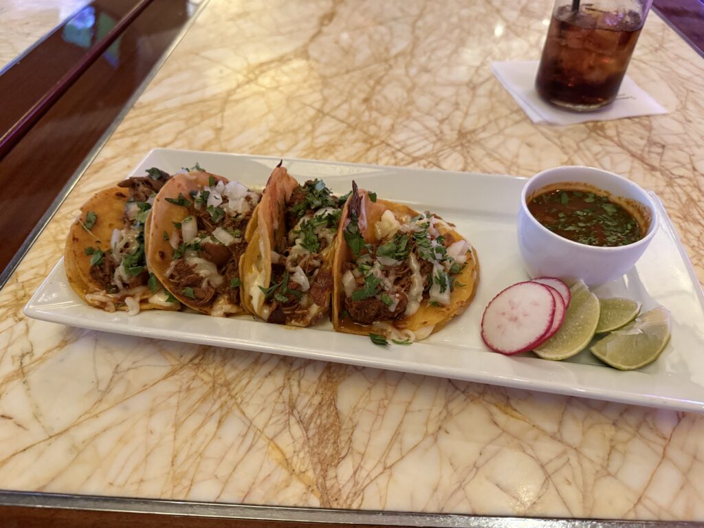 4 tacos lined up on a rectangular plate next to lime and radish slices. 
