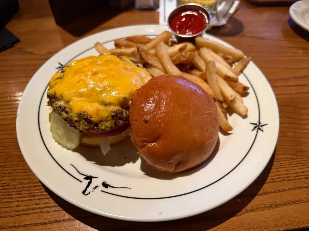 A cheeseburger with the top bun removed sits on a round white plate next to a pile of french fries. 