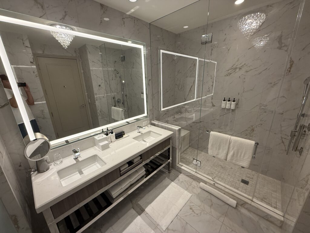 An illuminated mirror over a dual sink vanity with a glass enclosed shower in the background.