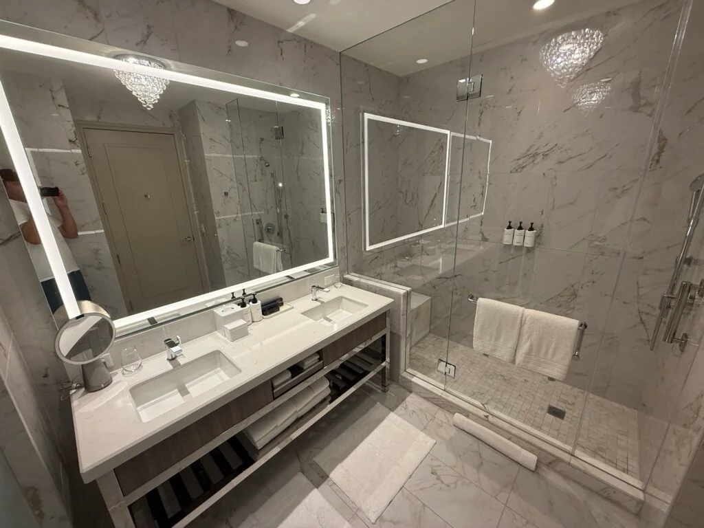 An illuminated mirror over a dual sink vanity with a glass enclosed shower in the background. 