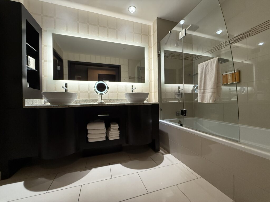 A dual sink vanity, with an backlit mirror above it and a shower to the right.