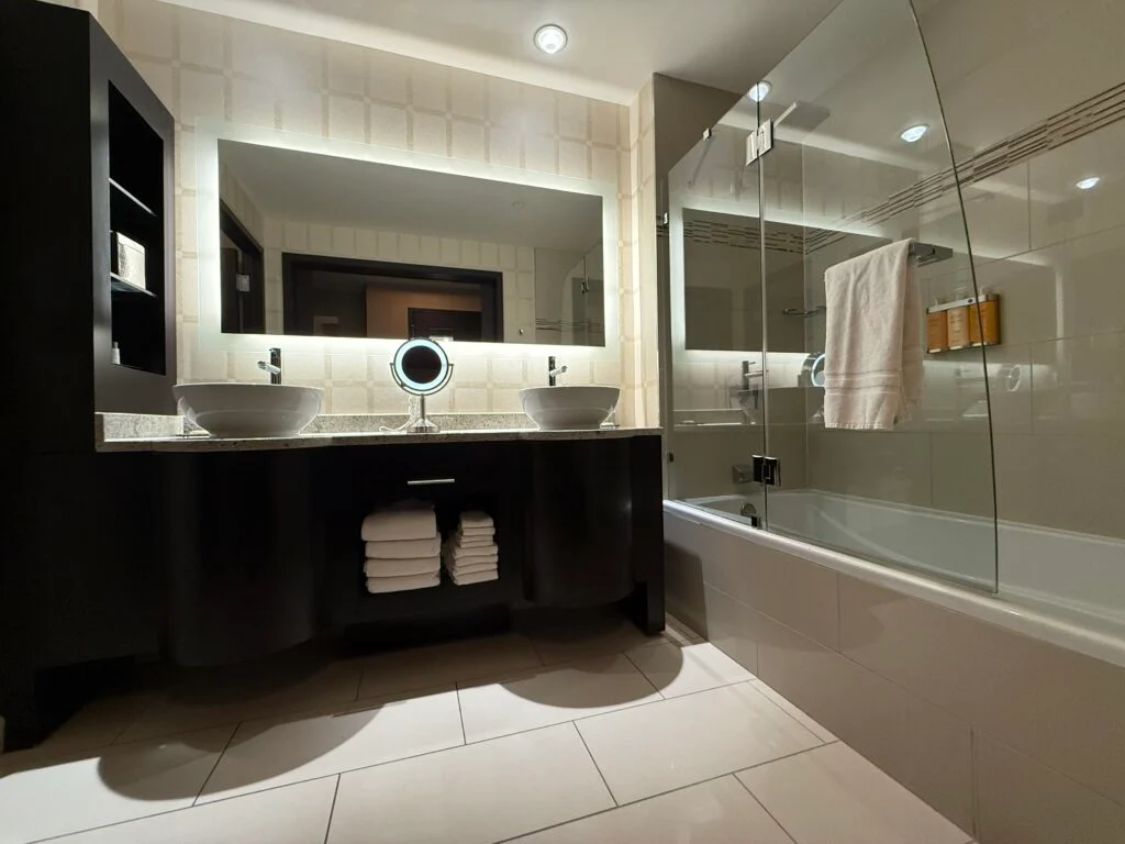 A dual sink vanity, with an backlit mirror above it and a shower to the right. 