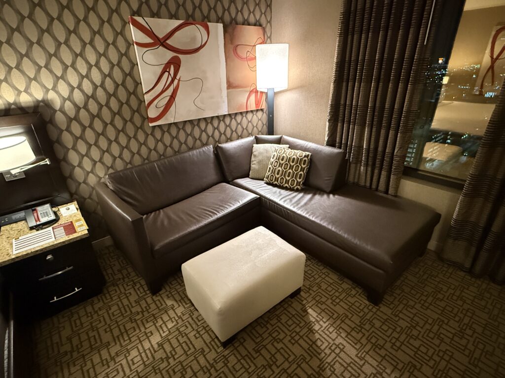 A brown, L shaped couch with a lamp behind it and a white ottoman in front.