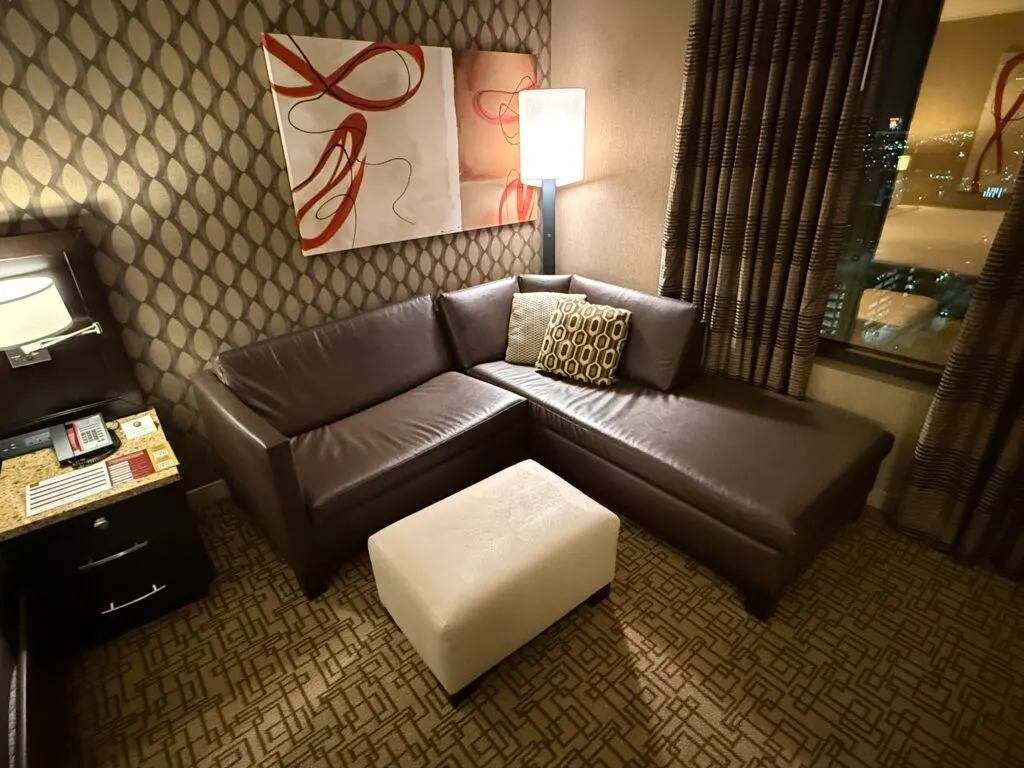 A brown, L shaped couch with a lamp behind it and a white ottoman in front. 