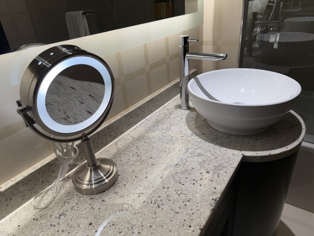 An illuminated makeup mirror on the counter, with a bowl sink in the background.