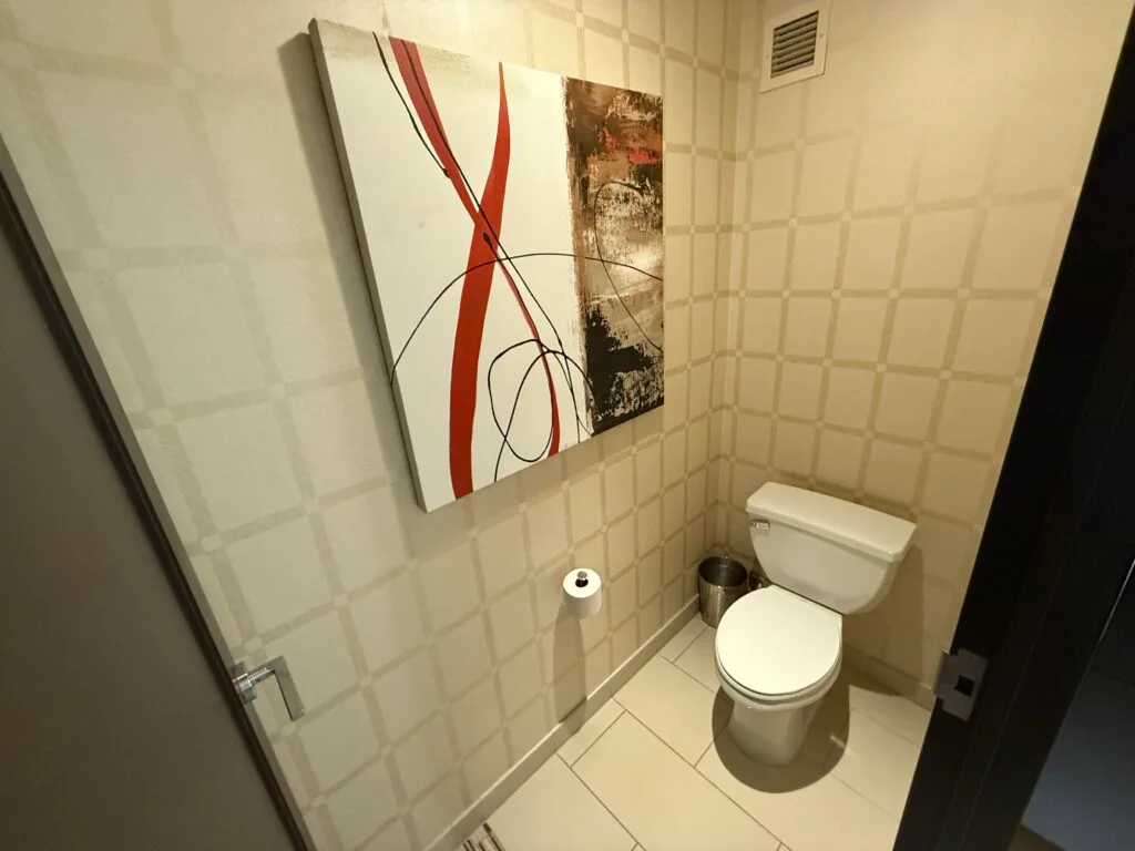 A toilet in a small room off the main bathroom with a piece of "swirly" art hanging above it. 