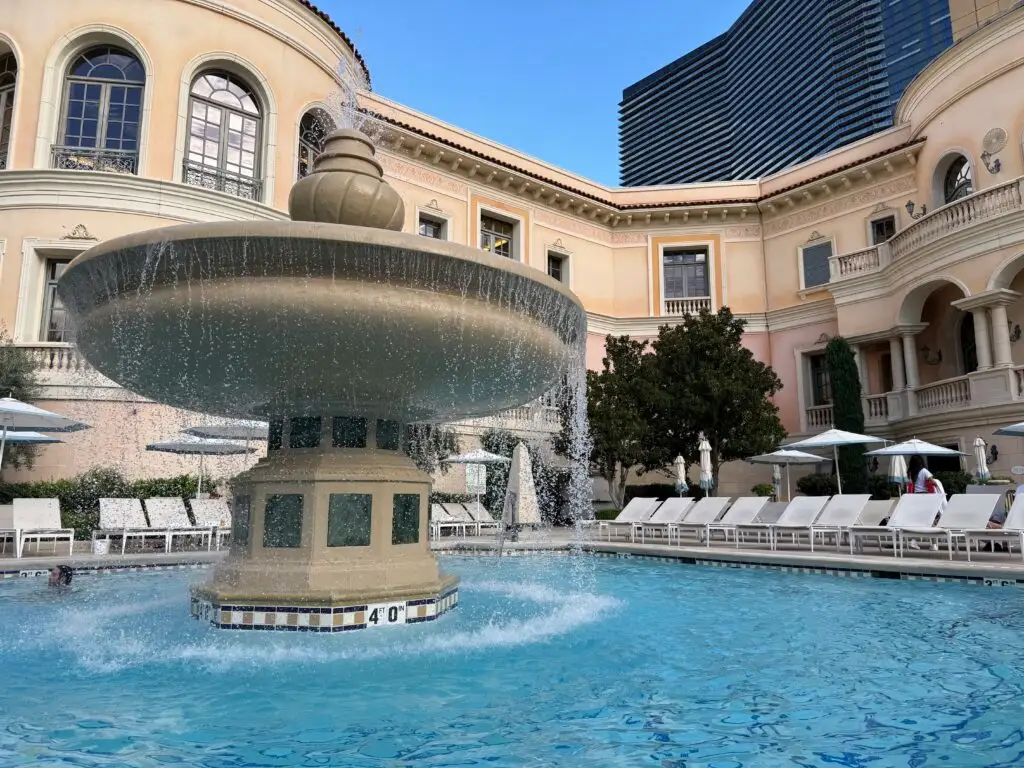 A large circular fountian in the middle of a pool, with The Cosmopolitan's htoel tower looking on in the background. 