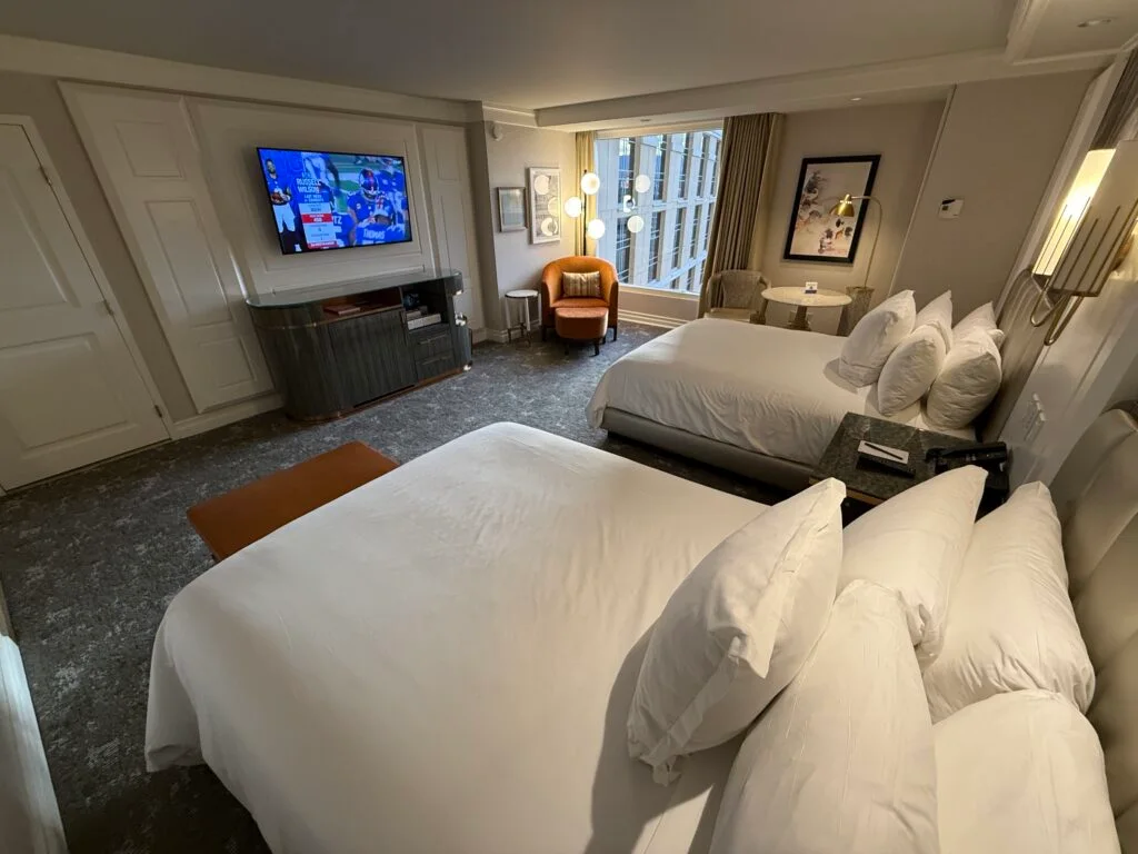 An angle of the room that shows the 2 queen beds pointed at the TV with the window and chairs in the background.